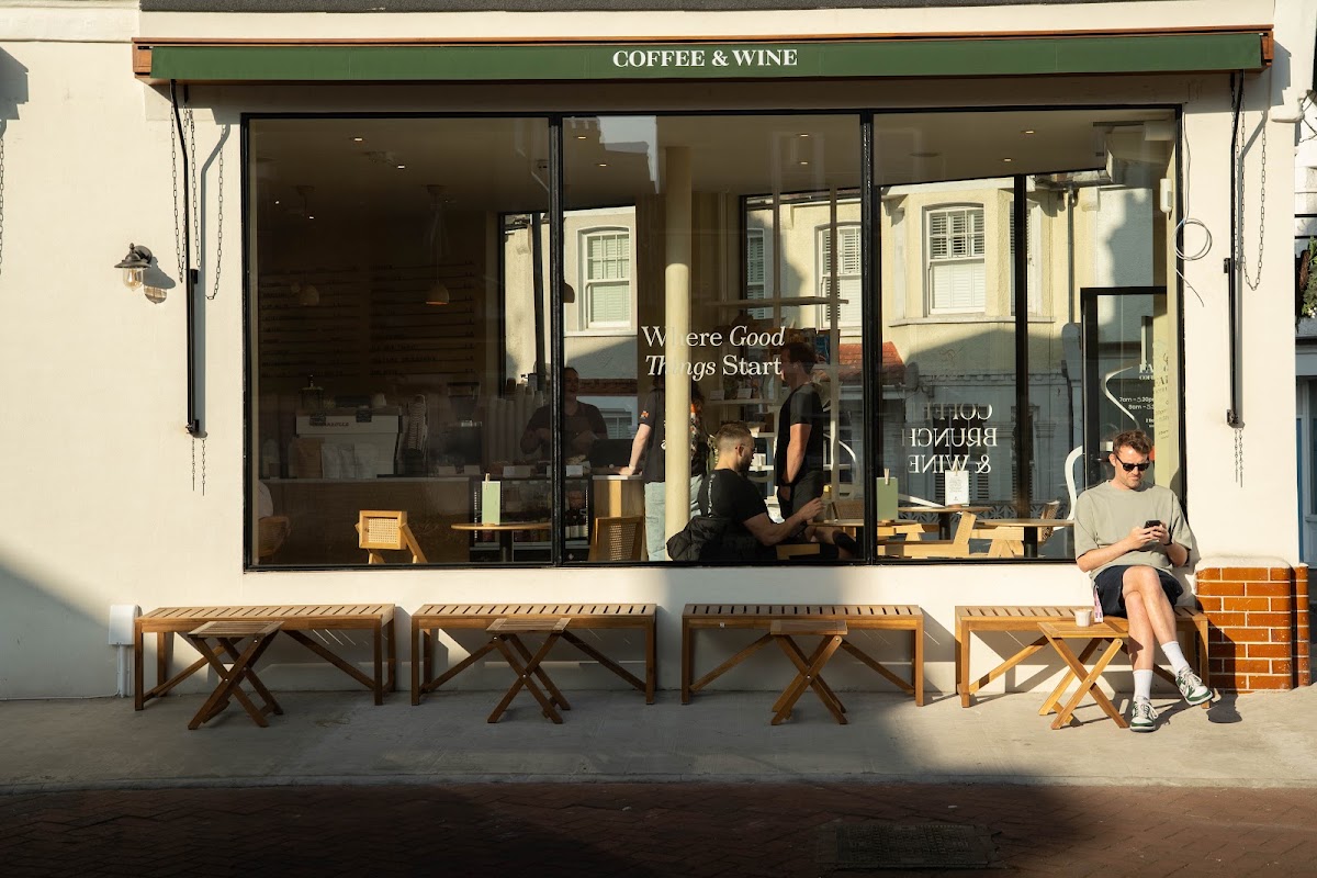 The front of Farine in late afternoon sun: a dark green awning lettered COFFEE & WINE above a wide black-framed window, with timber benches and folding tables along the pavement and customers sitting outside.