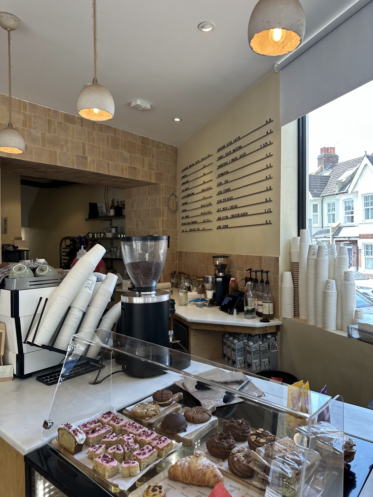 The counter inside Farine: a glass case of iced cakes, cookies and croissants, a marble top with coffee grinders, stacks of branded takeaway cups, tan tiled walls, concrete pendant lamps and the drinks board on the wall.